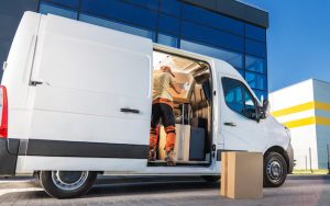 A courier loads cardboard boxes into a white van through the side door outside a warehouse building. It illustrates what shapes courier pricing UK, from handling and loading to choosing the right vehicle for the job.